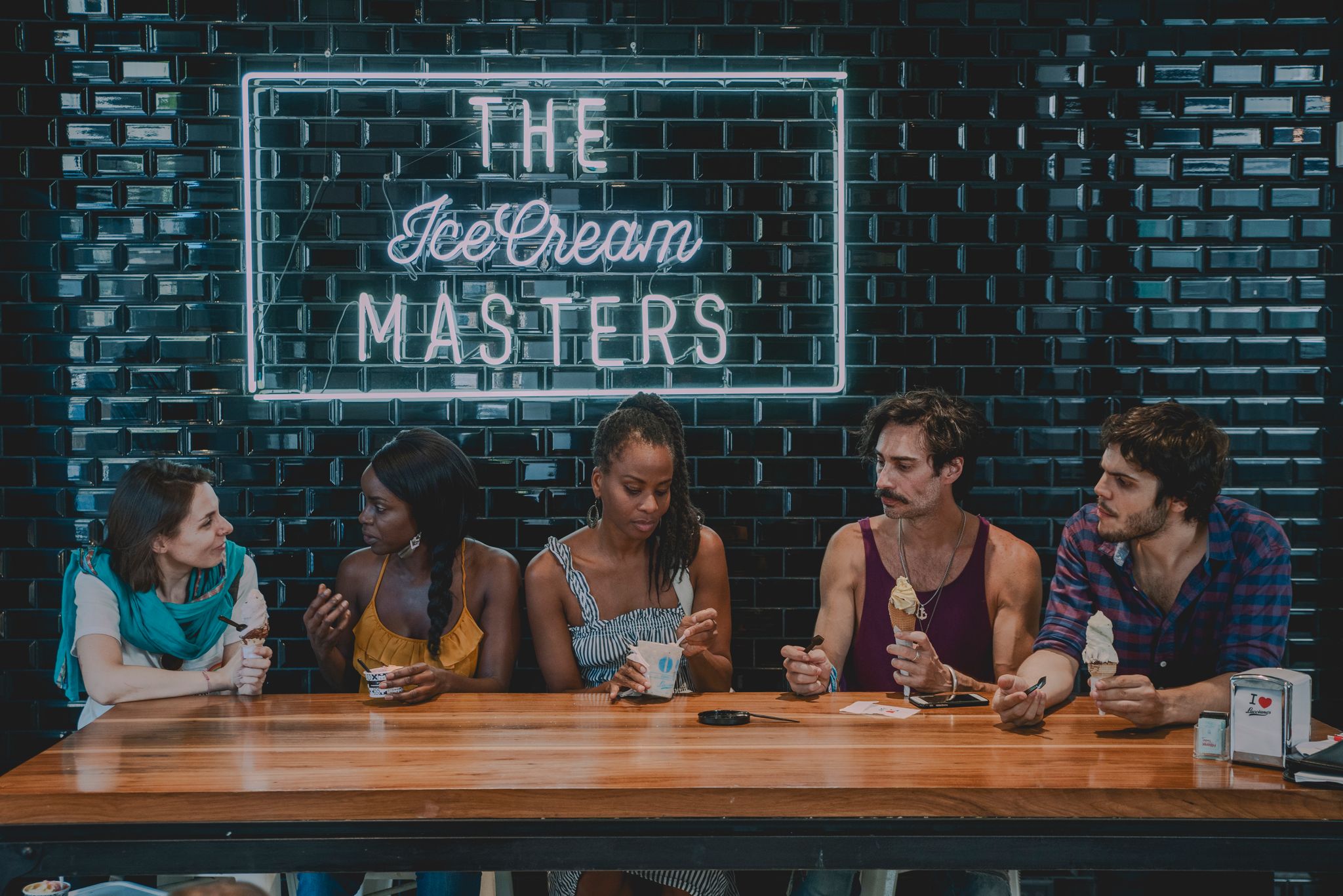 5 people are sitting at a wooden table. The first is a white woman with short brown hair. She's wearing turquoise scarf, a white shirt and has her arms folded. Beside her is a Black woman with long straight hair eating ice cream. She's wearing an orange tank top. Beside her is a Black woman with her braids in a pony tail. She's wearing a black and white striped dress and is eating ice cream. Beside her is a Latino man with a man bun and a maroon tank top eating ice cream. Beside him is another Latino man with dark hair and a mustache and beard eating ice cream. They are sitting in front of a deep turquose tiled wall, that has a fluorescent sign that says 'ice cream masters'. It is a fairly diverse customer base.