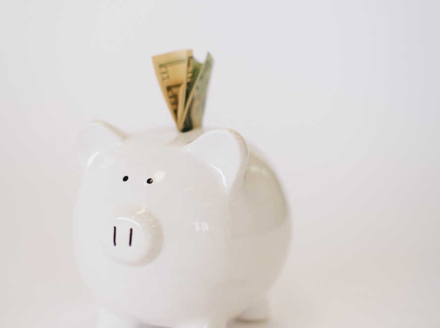 A white pig bank on a white background. US 20 dollar bills are folded up and stuck into the top part of the pig bank, and there are coins on a white background next to the pig bank.