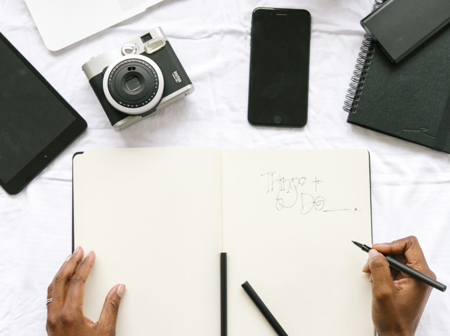 A complete work table, with a laptop, a watch, a smartphone, a tablet, a notepad, a camera and the hands of a person taking notes.