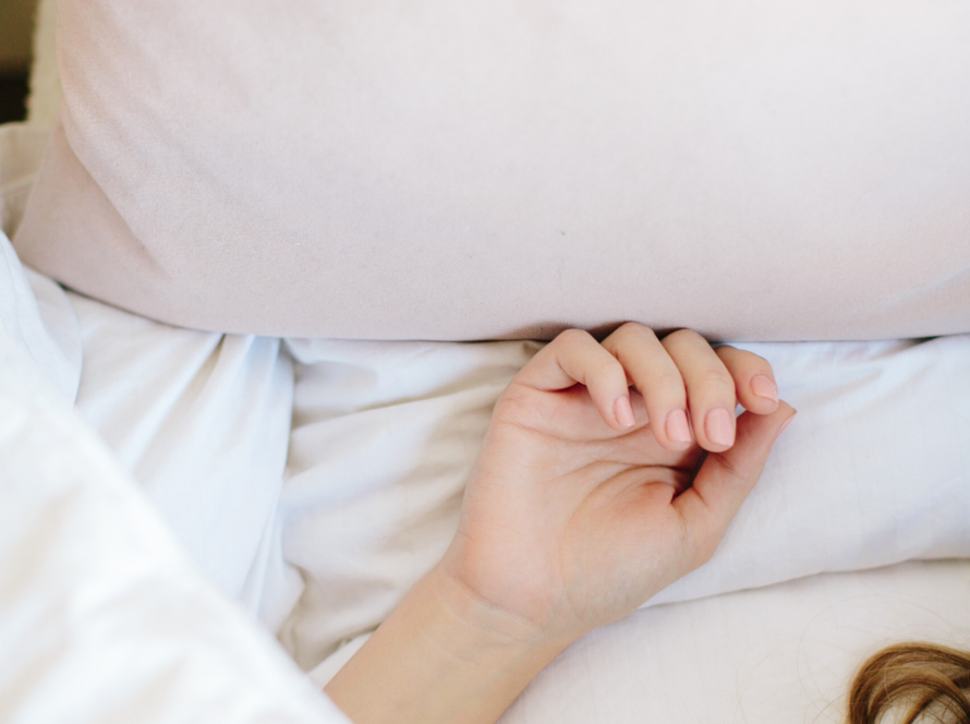 The image shows what appears to be a white woman with brown hair sleeping in a bed. She is under the white covers, laying on white sheets, with a beige pillow in a top of the image. We can see just a few locs of hair of the person in the lower right corner. It is meant to signify someone who is asleep, rather than 'woke' or awake