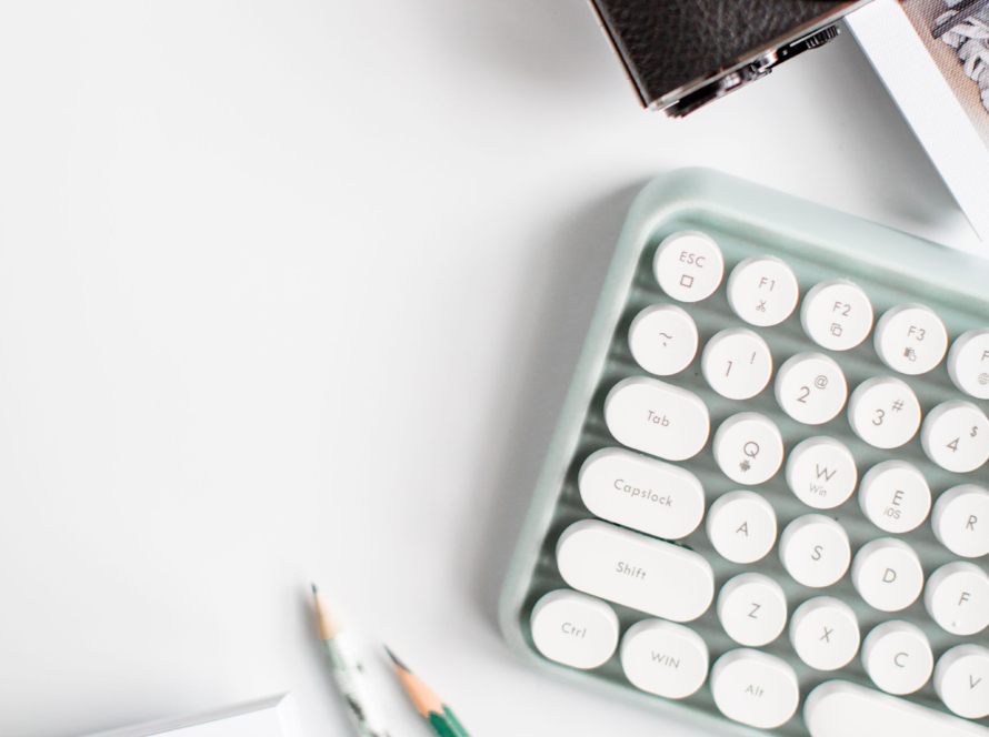 In the image we can see a camera, a PC keyboard, two black lead pencils and a notepad.