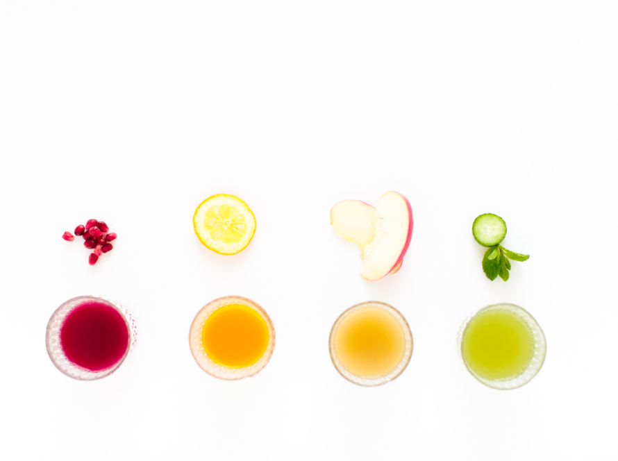 4 fruits with shot classes shown from above placed directly under the fruit. Each fruit (or piece of it) is a vibrant different color. The first are ppomogranites in a vibrant red. The second is a lemon slice in a vibrant yellow. The third are apple slices in a vibrant red. And the last is mint in a vibrant green.