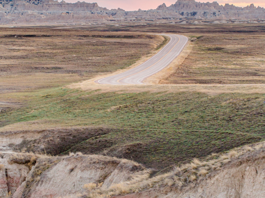 A curving road that leads to a city off in the distance. the terrain on either side of the road is empty, undeveloped even. There isn't a soul in sight. Behind the city in the distance are beautiful skies and lovely blue and pink horizon.