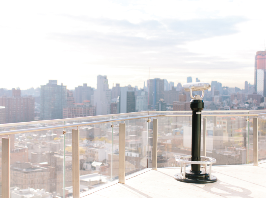 In the image we can see a telescope from a balcony and a panoramic view of the city