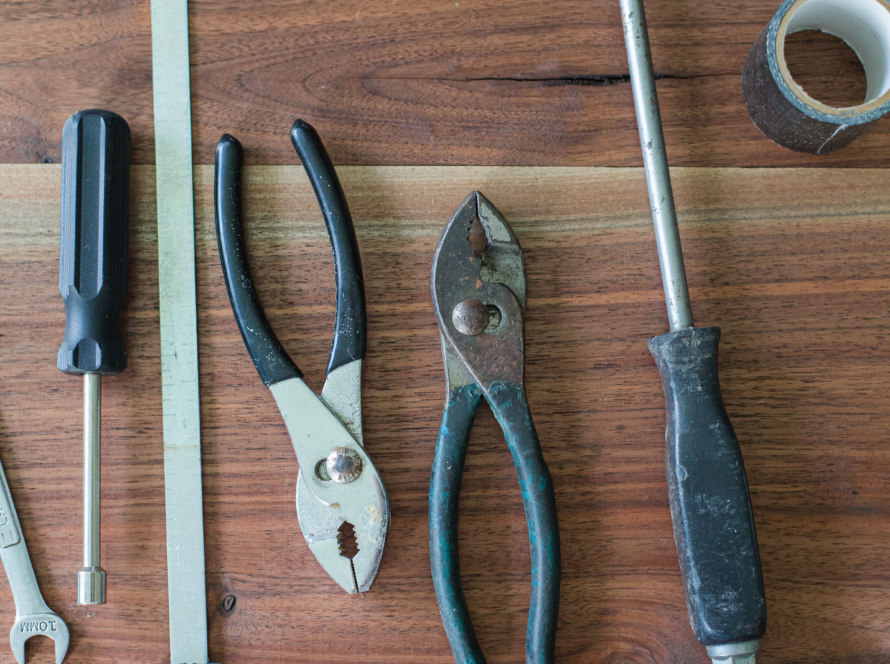 A wrench, a screwdrivers with a blue handle, a flat long rectangular piece of metal with a blue handle, a pair of pliers with a blue handle, another pair of pliers with a blue handle -- placed in the opposite direction, and another bigger screwdriver with a blue handle, and a roll of duct tape are place on a wood table -- with the camera looking at the tools from overhead.