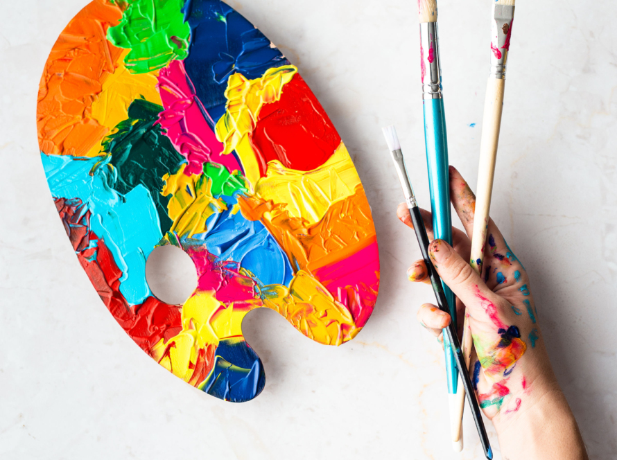 In the image, we can see a watercolor palette with many colors and a hand with three brushes