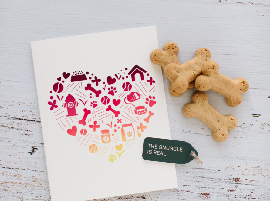 A white card is testing on an off white distressed table. On the card is a heart made up of red, purple, and orange images of items related to dogs, such as dog bones, hearts, a dog collar, a fire hydrant, a dog house, paw, dog food bowl, and grass. Next to the card are 4 brown dog bones, and there is a tag that says "the snuggle is real" in all caps. This is an abstract way of symbolizing brand inclusion -- by the inclusion of dogs.