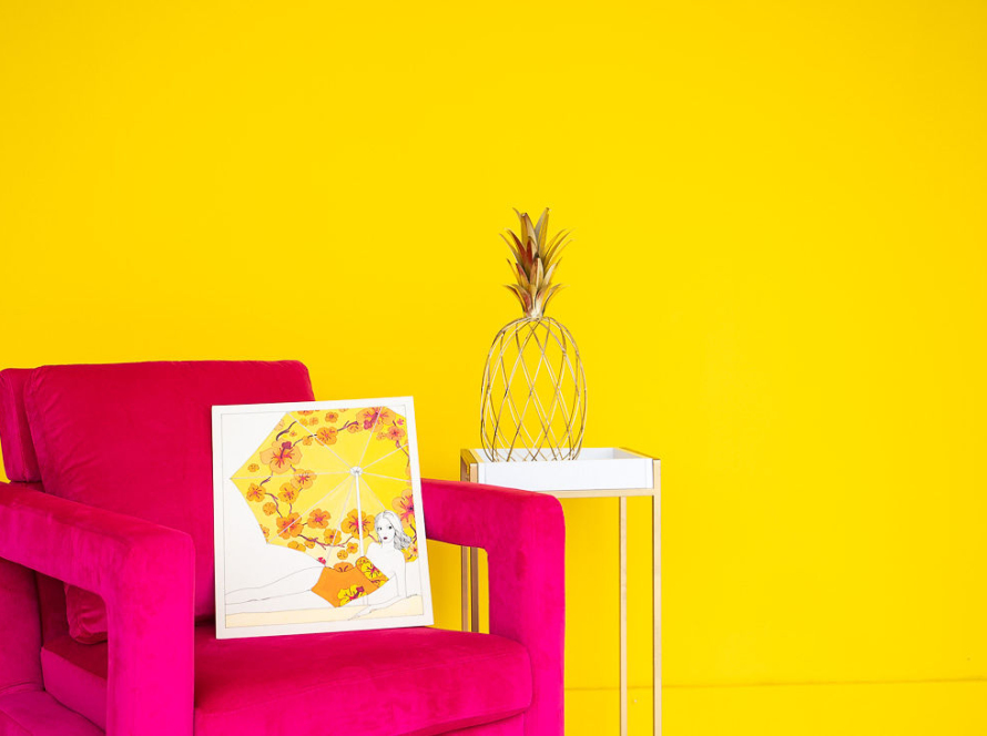 In the image, we can see a fuchsia armchair on a bright yellow background, a painting on top of it, and a pineapple-shaped ornament.