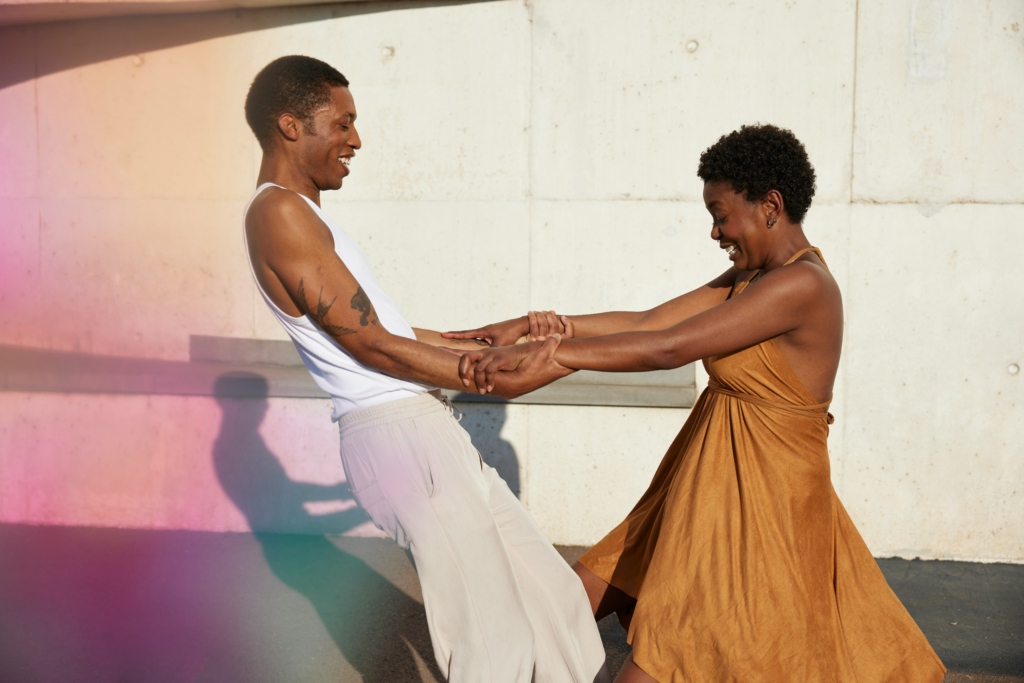 Am image showcasing a Black man and woman holding each others arms. The Black man on the left, wearing a white tank with khacki pants. He has short black hair, and is smiling. He is leaning back from the woman as if he is trusting her to hold him up. The woman is on the right, and has her arms extended holding the mans arms. She is smiling and looking down, and wearing a brown, burnt orange flowy sundress. She has a short afro. Image Symbolizes trust in marketing.