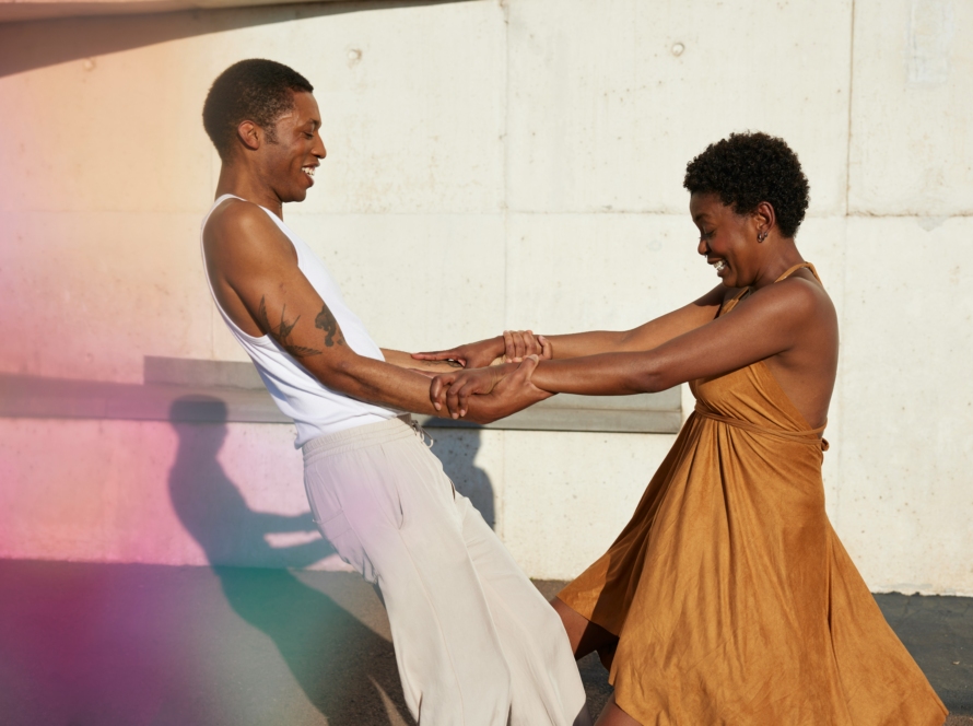 Am image showcasing a Black man and woman holding each others arms. The Black man on the left, wearing a white tank with khacki pants. He has short black hair, and is smiling. He is leaning back from the woman as if he is trusting her to hold him up. The woman is on the right, and has her arms extended holding the mans arms. She is smiling and looking down, and wearing a brown, burnt orange flowy sundress. She has a short afro. Image Symbolizes trust in marketing.