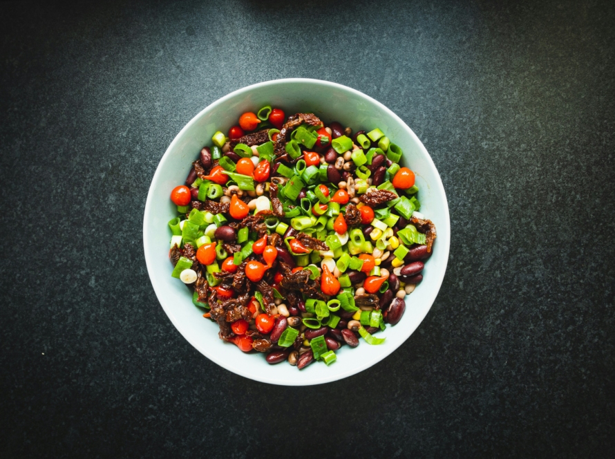 On a black lit background, there is a white bowl in the center filled with a colorful bean base salad. It also has cherry tomatoes mixed in and green onions.