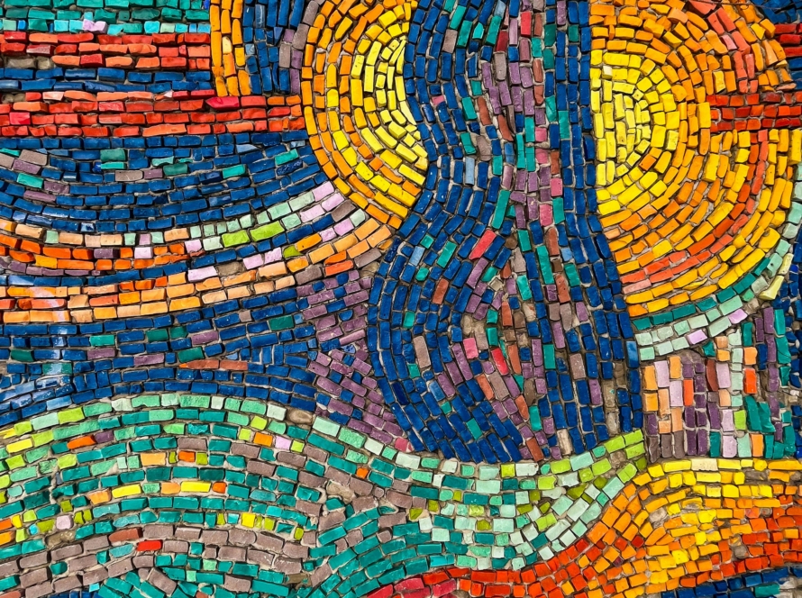 Close-up of a handmade mosaic built from hundreds of small, irregular tiles. Two sun-like circles of yellow and orange tiles glow near the top, split by a flowing river of deep blue. Wavy rows of teal, coral, purple, pink and orange ripple across the rest of the piece, so it feels like it's in motion. Each tile is a different shade, but together they form one picture.