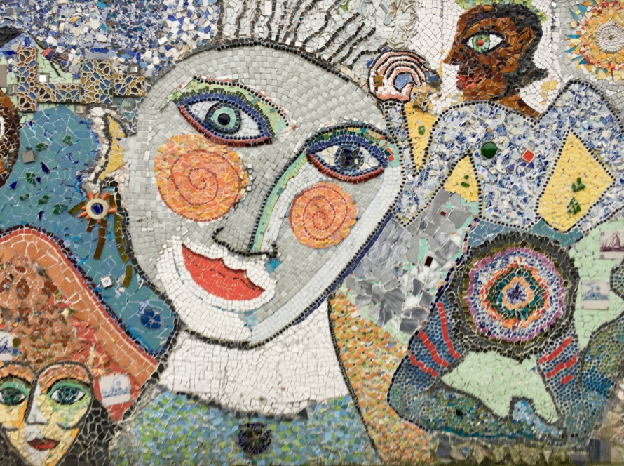 Colorful mosaic mural made of tile and broken ceramic, showing several faces in different colors and styles, including a large stylized face with spiral orange cheeks, surrounded by patterns and shapes.
